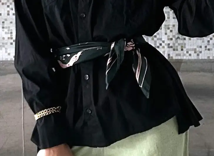 Detalle: un pañuelo estampado anudado sobre el cinturón de una camisa negra, y una pulsera de cadena dorada.